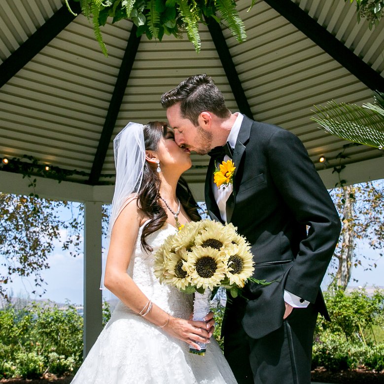 Bride and groom kissing under a gazebo, holding a bouquet of sunflowers.