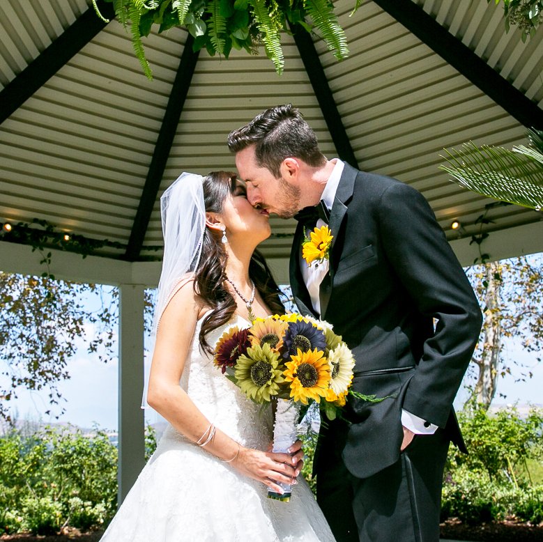 Bride and groom kissing under a gazebo, holding a bouquet of sunflowers and dark red flowers.