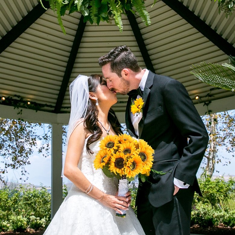 Bride and groom kissing under a gazebo, bride holds a bouquet of sunflowers