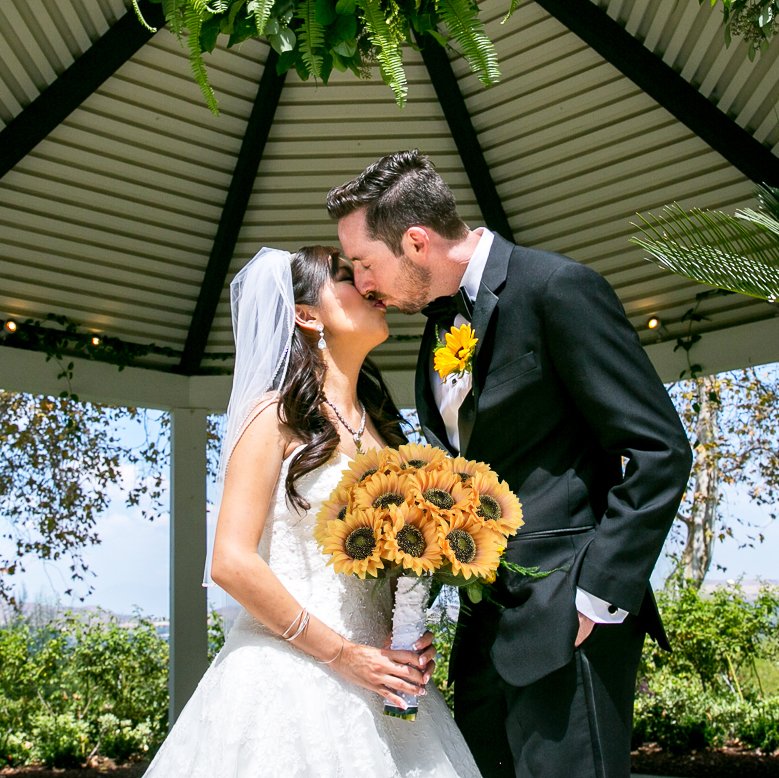 Bride and groom kissing under a gazebo, holding a bouquet of silk sunflowers.