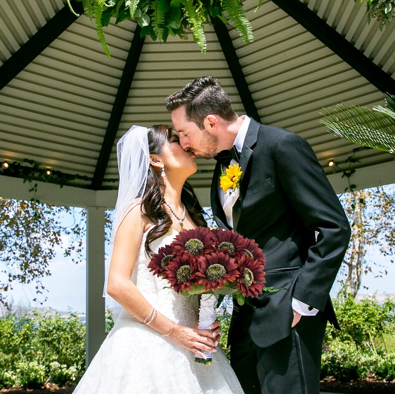 Bride and groom kissing under a gazebo, holding a bouquet of dark red sunflowers.