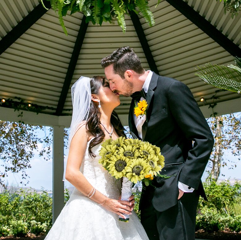 Bride and groom kissing under a gazebo, bride holds a bouquet of sunflowers.