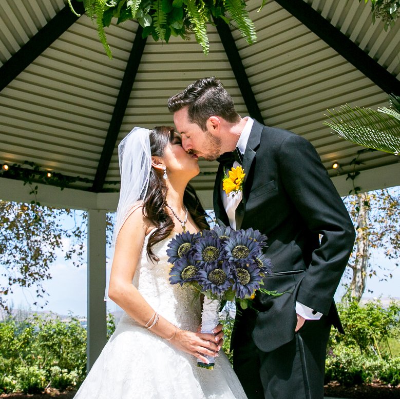 Bride and groom kissing under a gazebo, holding a bouquet of dark blue sunflowers.