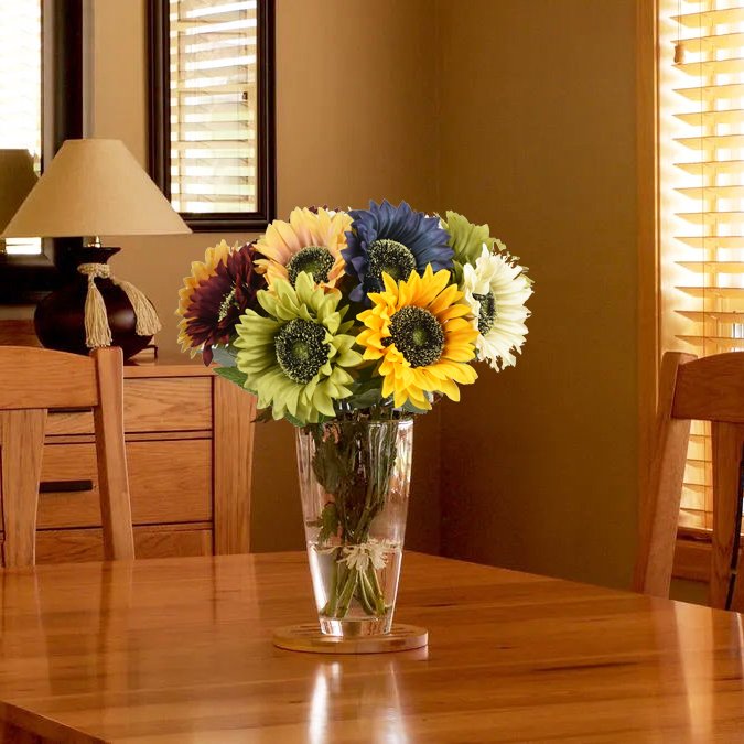 Bouquet of artificial sunflowers in various colors in a glass vase on a wooden table, with a lamp and chair in the background.