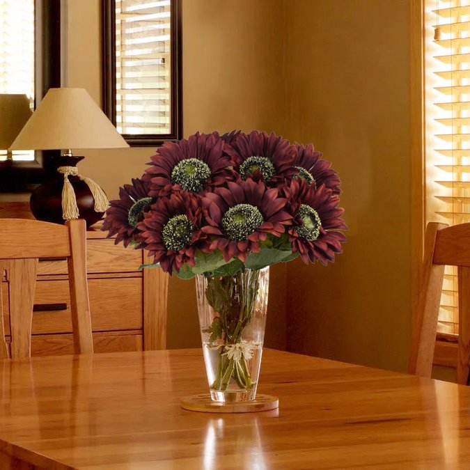 Bouquet of dark red silk sunflowers in a clear glass vase on a wooden table, with a lamp and window in the background.