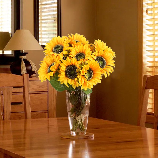 Bouquet of artificial sunflowers in a clear glass vase on a wooden table, with a lamp and window in the background.