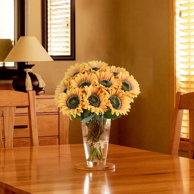 Bouquet of silk sunflowers in a clear glass vase on a wooden table, with a lamp and window in the background.
