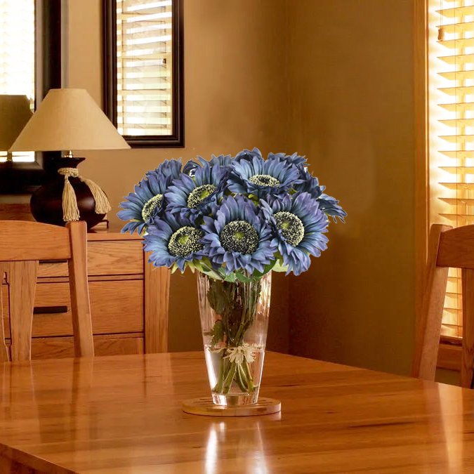 Bouquet of blue silk sunflowers in a clear glass vase on a wooden table, with a lamp and window in the background.