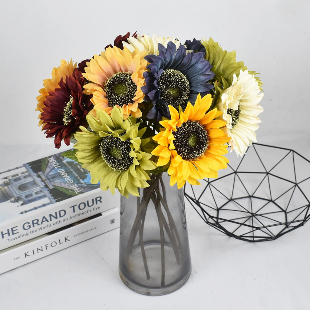 Bouquet of artificial sunflowers in various colors in a grey vase, next to books and a wire basket.