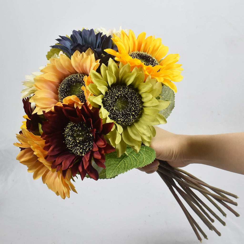 Hand holding a bouquet of artificial sunflowers in various colors including yellow, orange, burgundy, and dark blue, with green leaves.