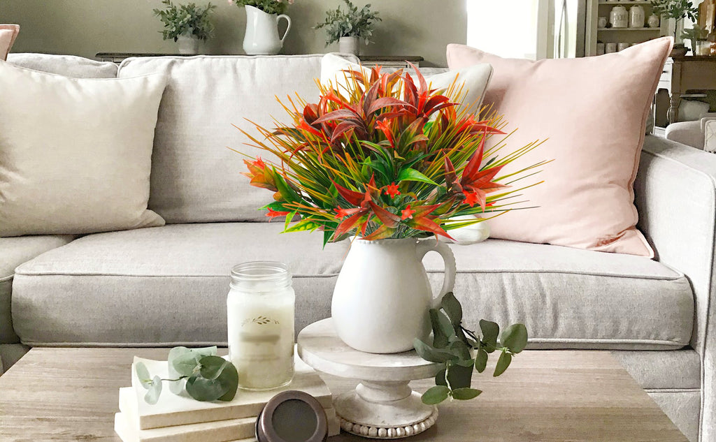 Artificial morning glory flowers in a white pitcher on a coffee table with a candle and books.