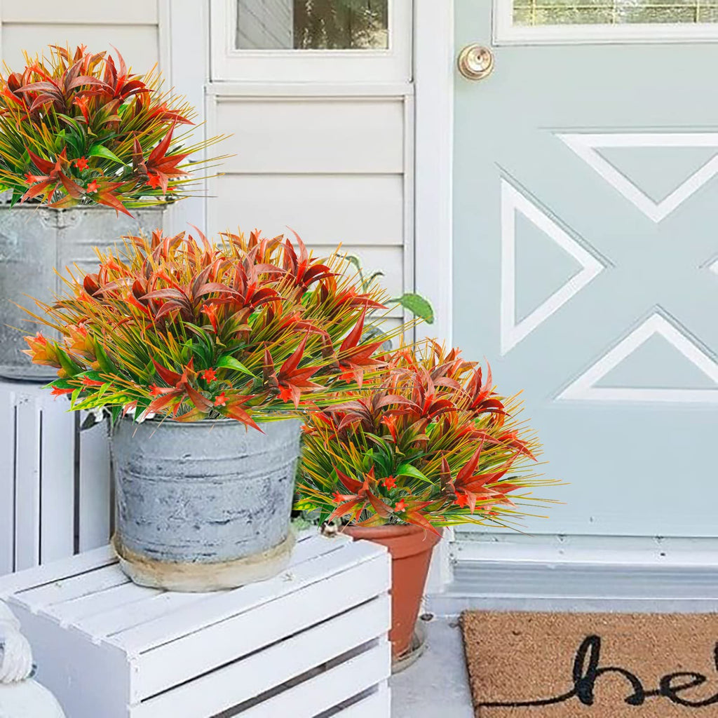 Three bunches of artificial red and orange morning glory flowers with green and yellow grass in pots on a white wooden crate by a light blue door.