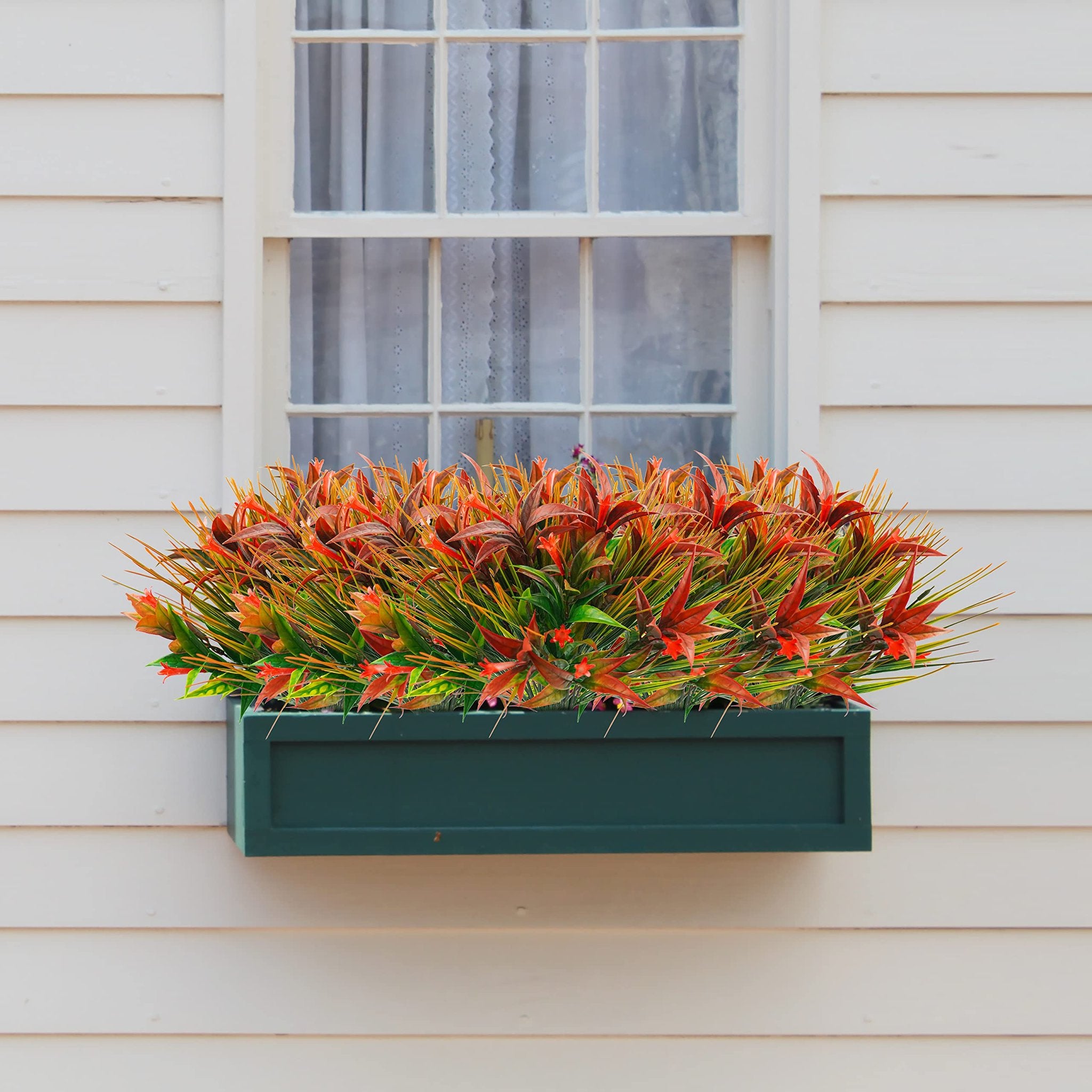 Artificial morning glory flowers and orange grass in a dark green planter box mounted on a white clapboard wall below a window.