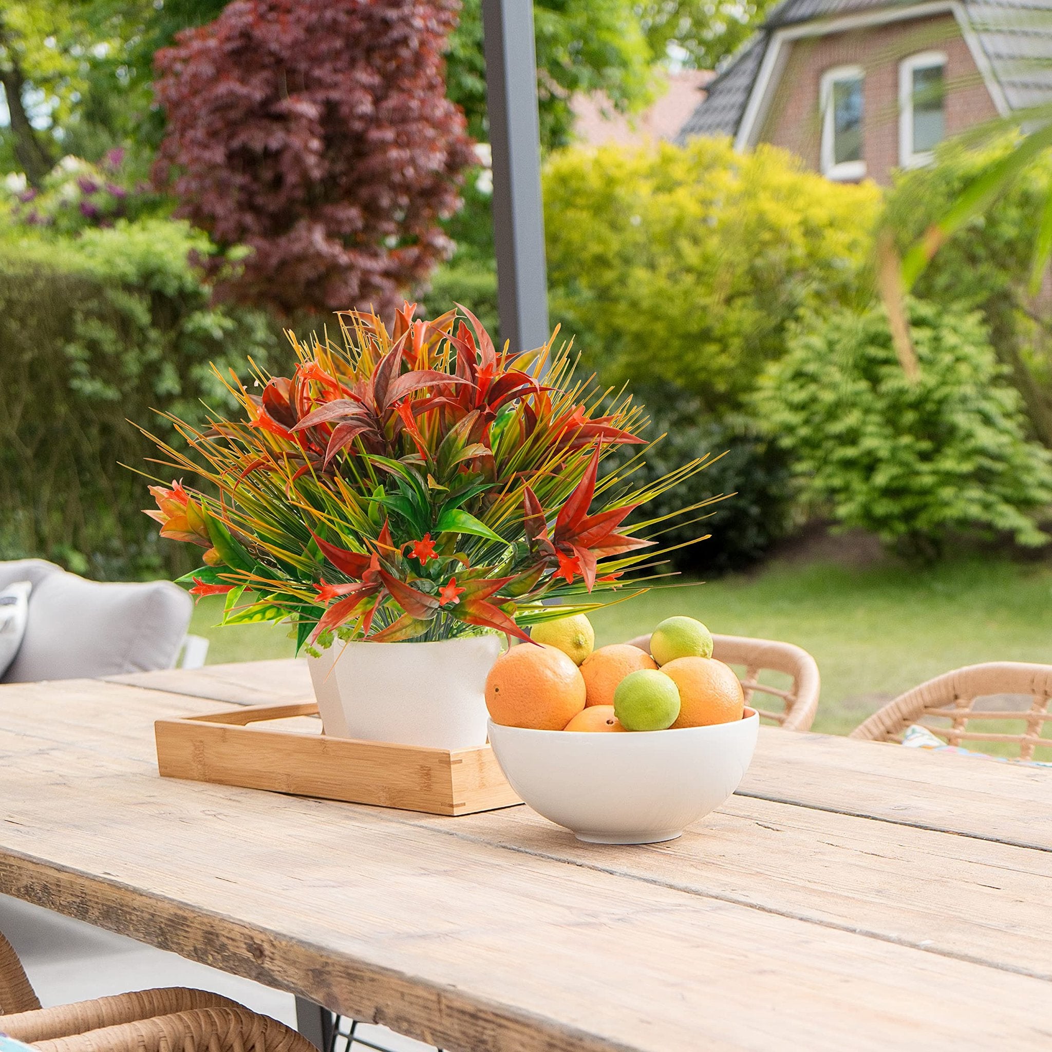 Artificial red and orange foliage in a white pot on a wooden table with a bowl of fruit