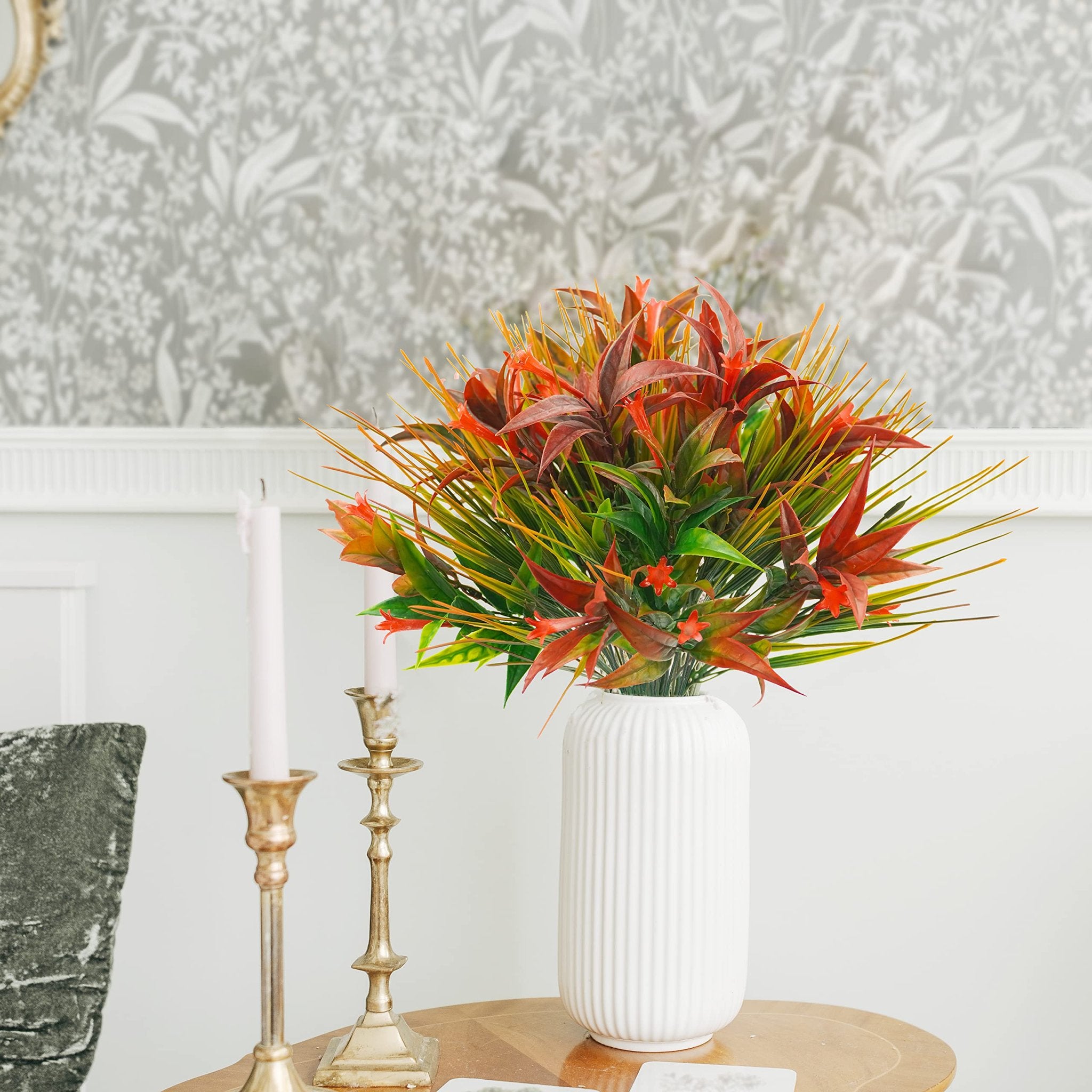Artificial red and orange morning glory flowers with green and yellow grass in a white vase on a table.