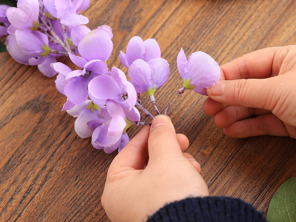 Hands arranging purple artificial wisteria flowers on a wooden surface