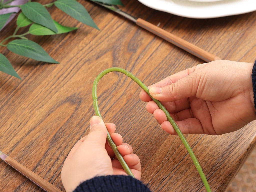 Hands bending a green artificial vine stem on a wooden table, with green leaves in the background.