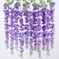 Six strands of artificial purple wisteria flowers with green leaves hanging against a white background.