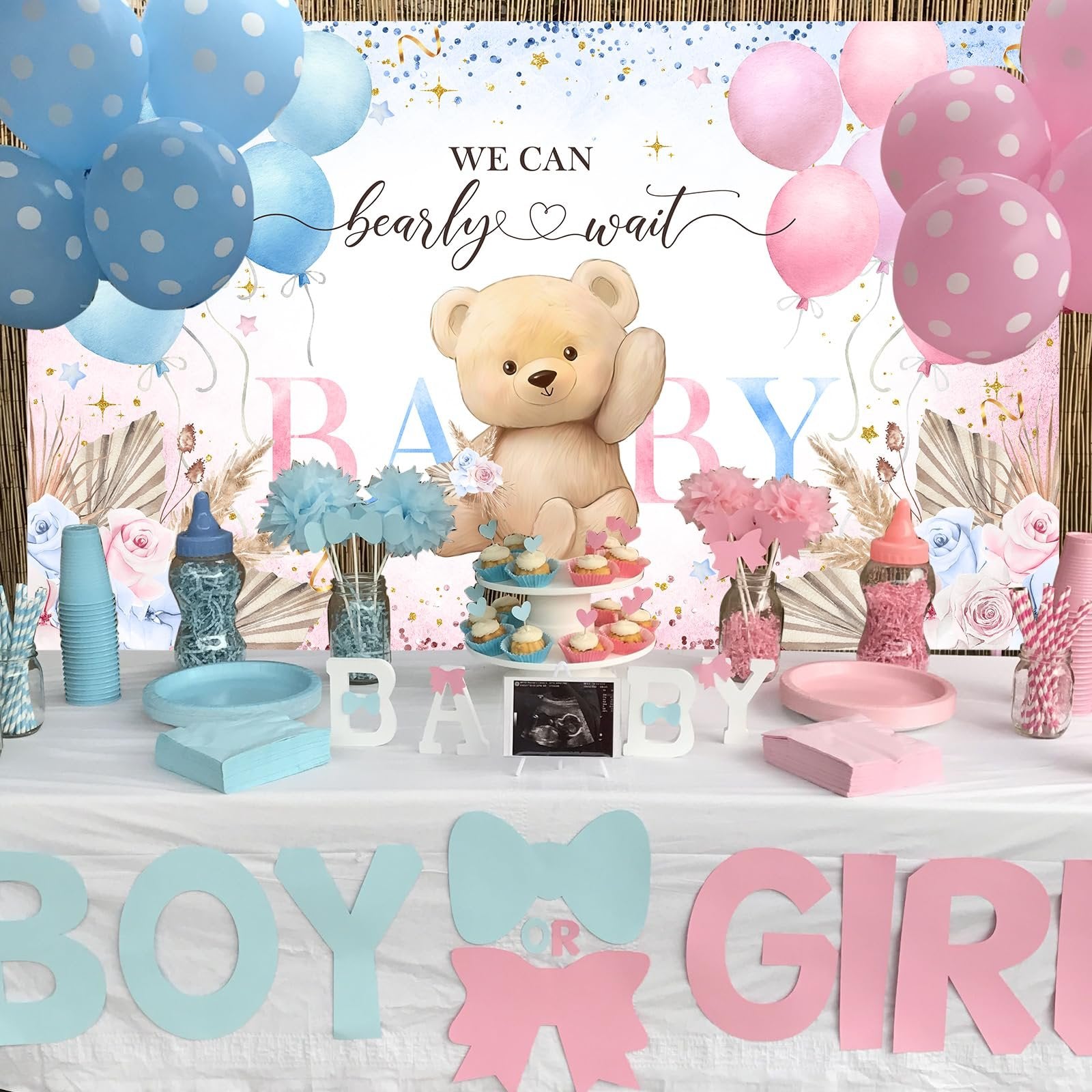 Gender reveal party table setup with blue and pink balloons, a teddy bear, cupcakes, and 'Boy or Girl' banner.
