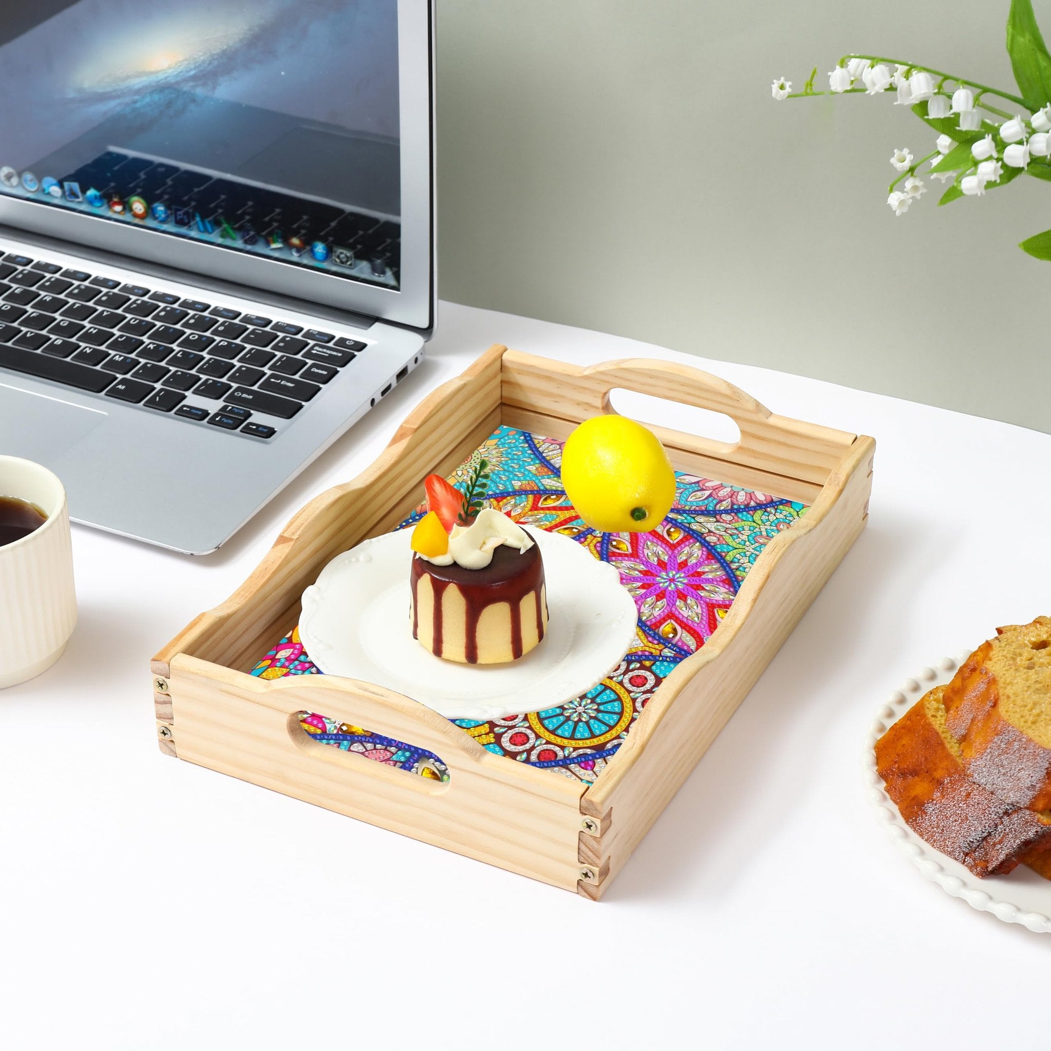 Wooden serving tray with a colorful mandala pattern insert, holding a small cake and a lemon, next to a laptop and coffee.
