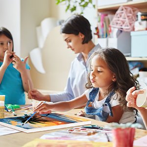 Children painting a framed canvas with a brush, guided by an adult in a classroom setting.