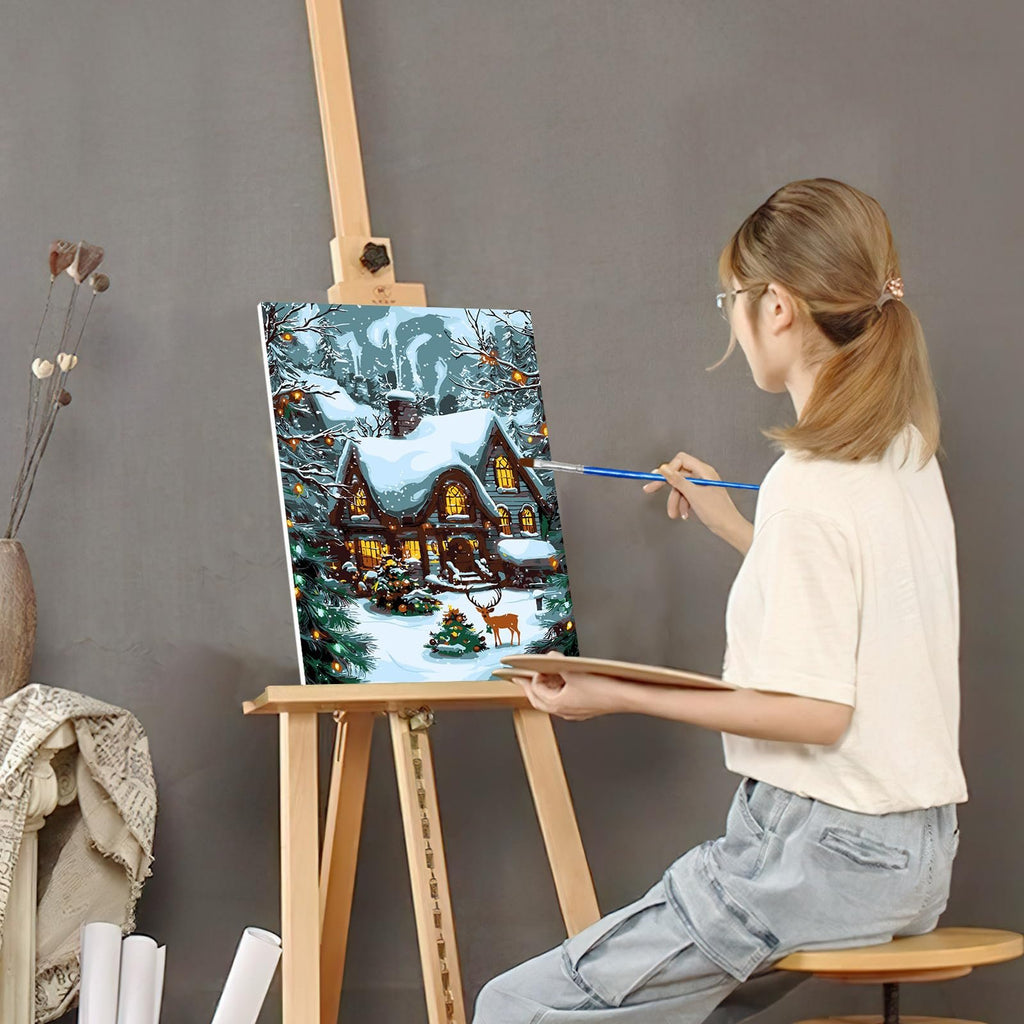 Woman painting a Christmas house scene on canvas on an easel, holding a palette and brush
