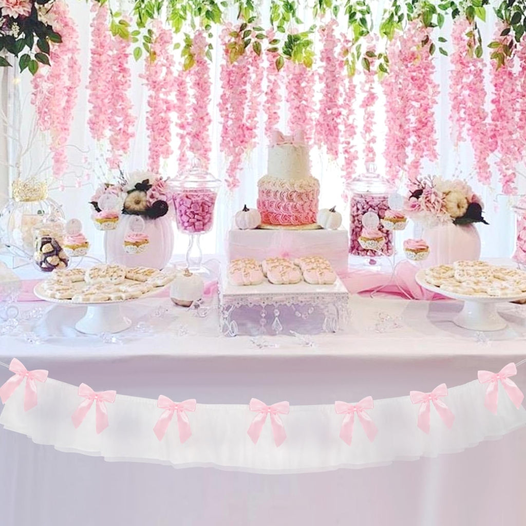 White ruffled banner with pink bows hanging above a dessert table with a cake and cookies