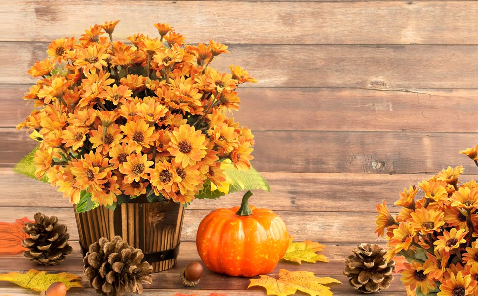 Artificial orange mums in a wooden bucket with a pumpkin, pinecones, and autumn leaves against a wood plank background