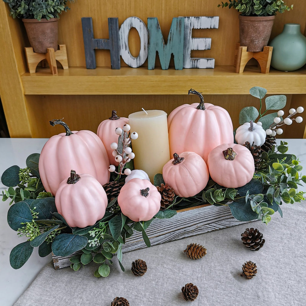 Assorted pink and white foam pumpkins arranged in a wooden crate with greenery and pinecones, a candle in the center.