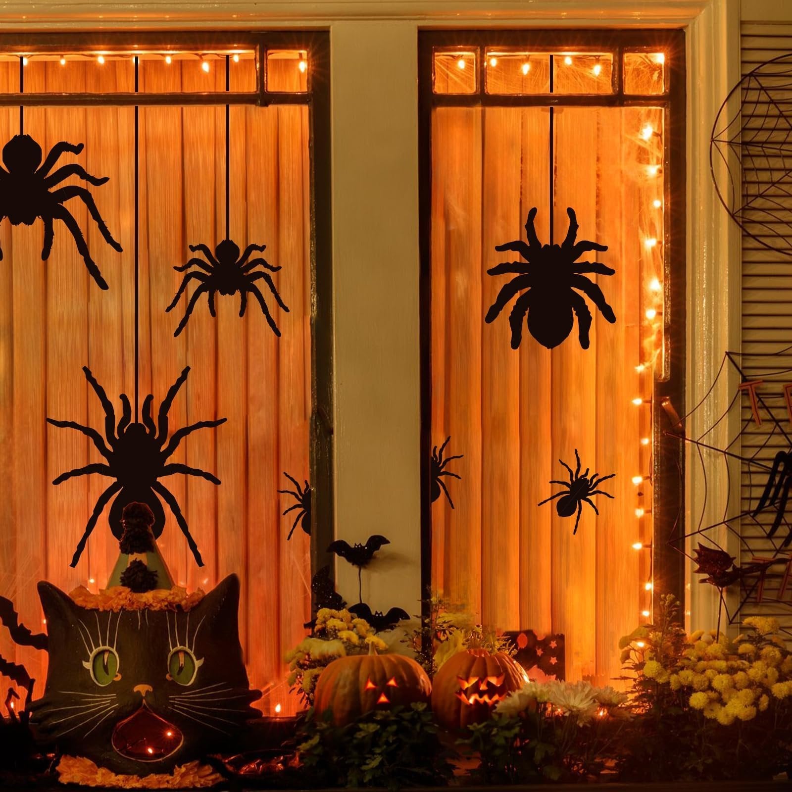 Halloween window decor with black spider silhouettes hanging in front of orange blinds, illuminated by string lights.