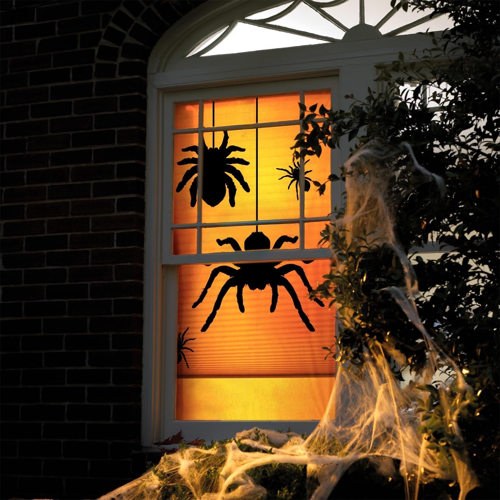 Halloween window decorated with large black spider silhouette clings and fake cobwebs on a brick house exterior.