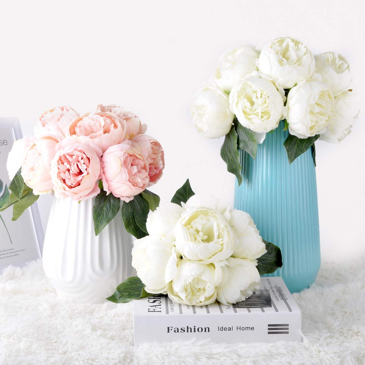 Bouquets of pink and white artificial peonies in vases and on a book, with green leaves.