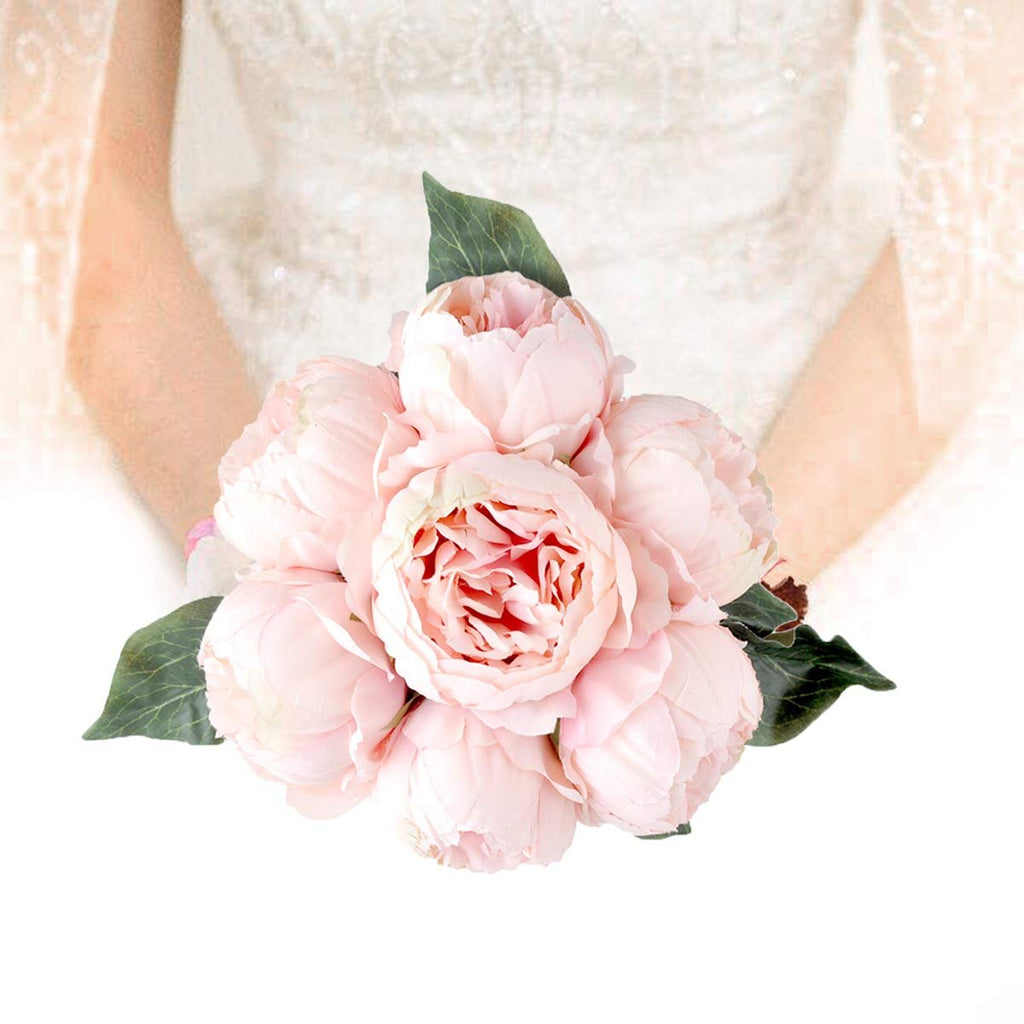 Bouquet of seven light pink peonies with green leaves held by a person in a white lace dress