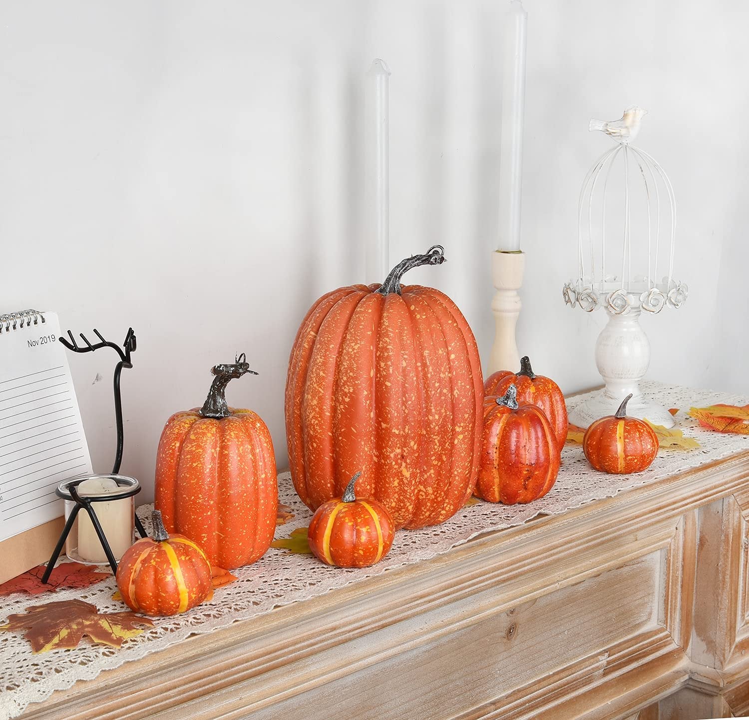 Assortment of seven artificial orange pumpkins of varying sizes arranged on a lace runner with faux fall leaves and two white candles.