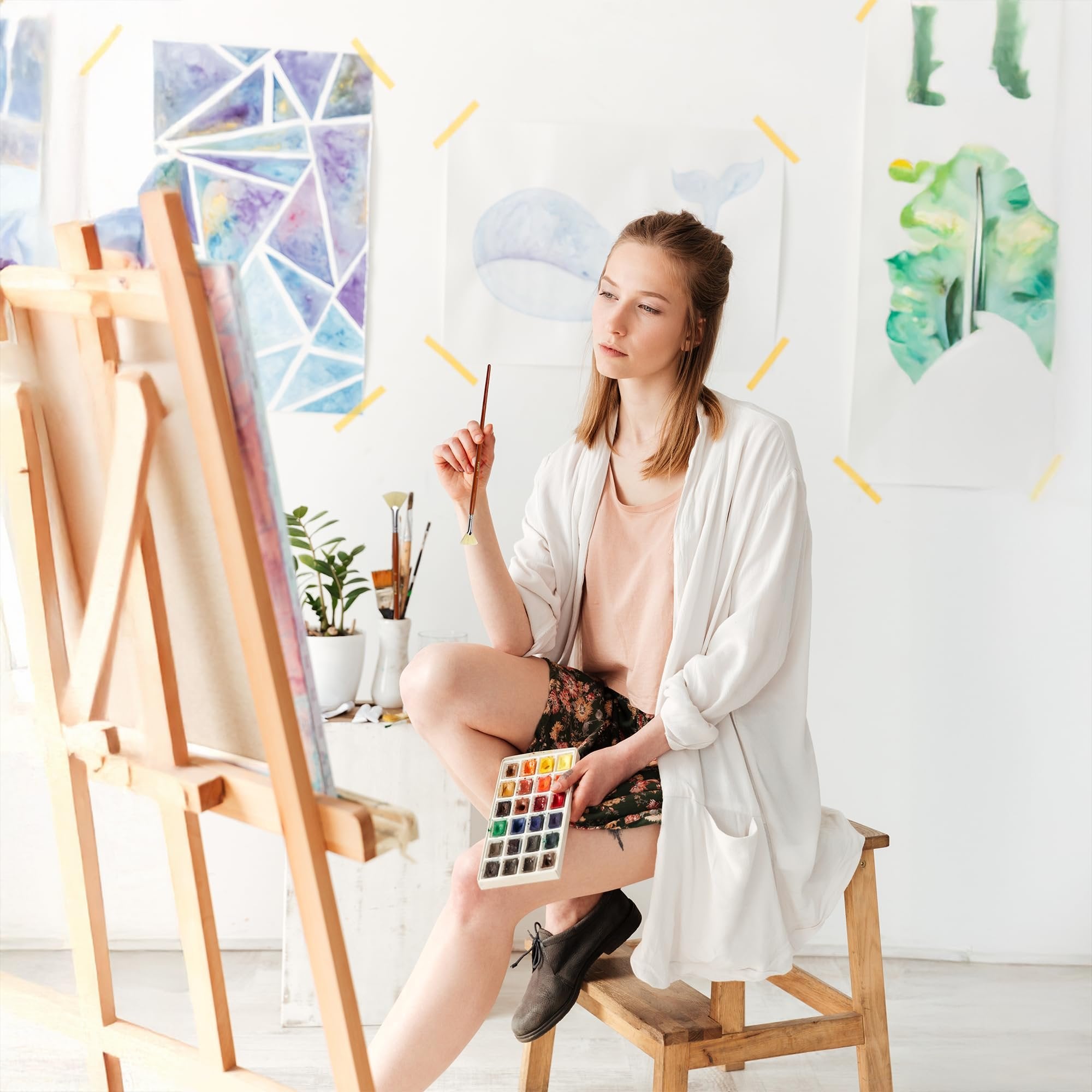 Young woman artist sitting on a stool in a studio holding a watercolor palette and a fan brush, looking at a canvas on an easel.