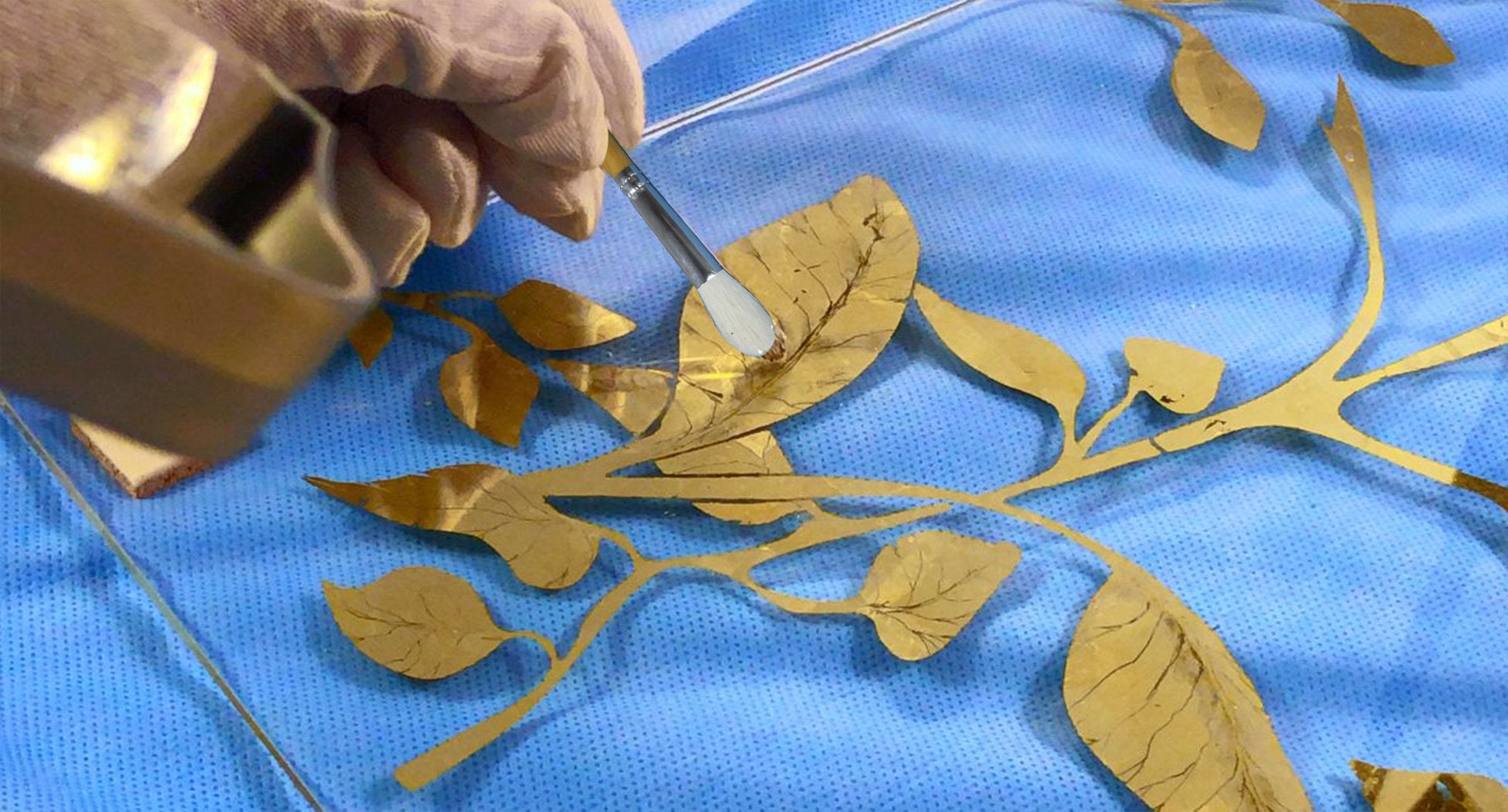Gloved hand uses a brush to apply gold leaf to a decorative branch on a blue textured surface.