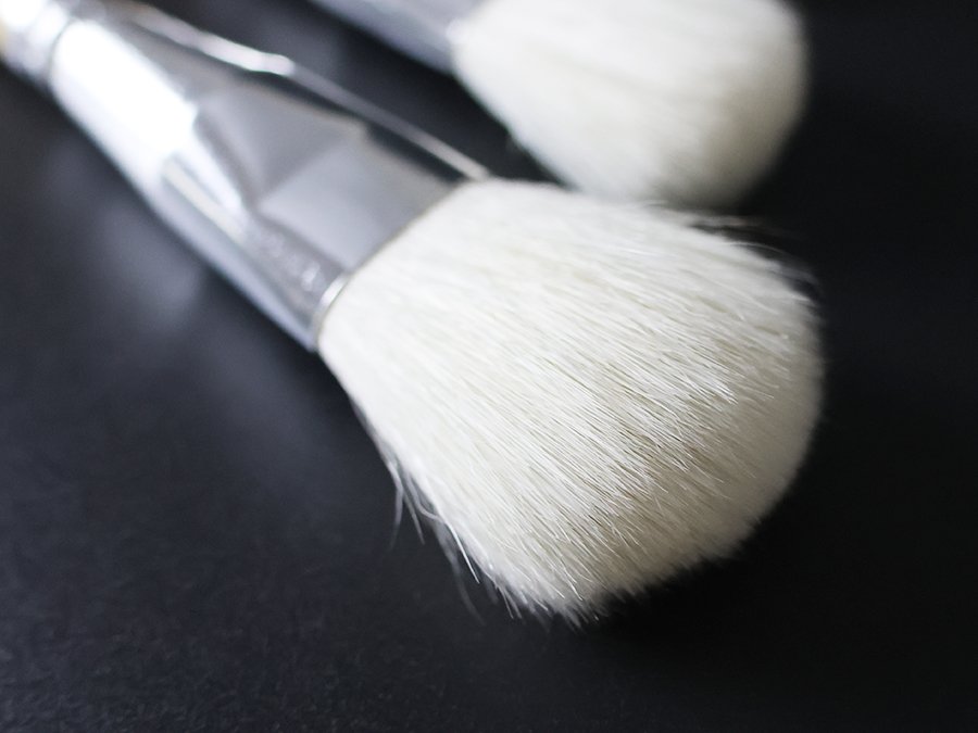 Close-up of two white goat hair gilding brushes with silver ferrules and birch wood handles on a black surface.