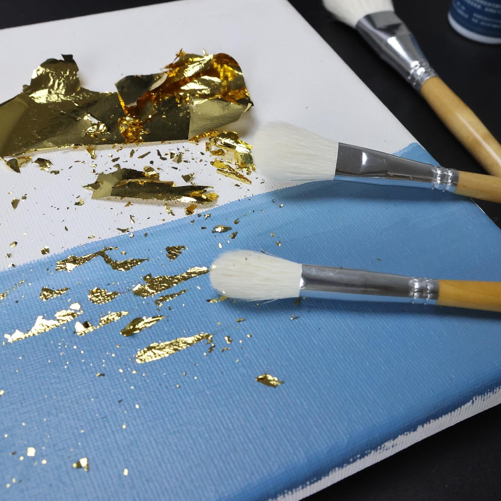 Gilding brushes applying gold leaf flakes to a blue and white canvas surface