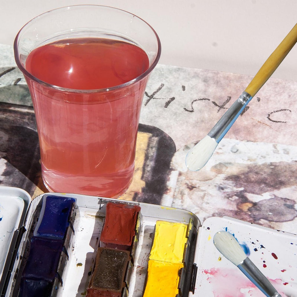 Watercolor paints, brushes, and a glass of pink liquid on a textured surface with artistic markings.