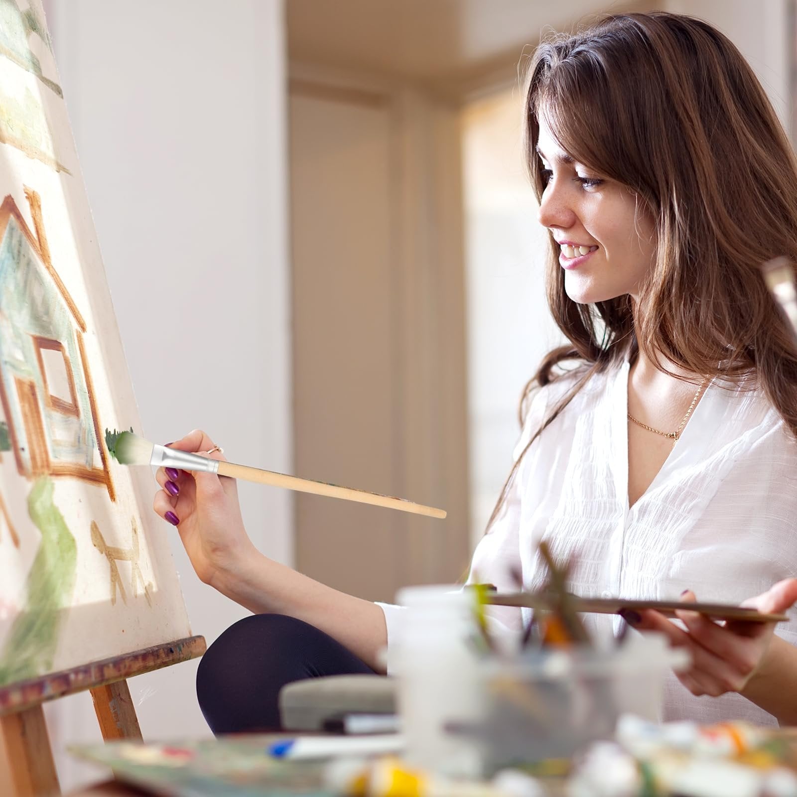 Young woman painting a house on a canvas with a brush, holding a palette