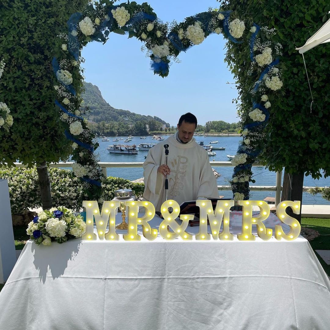 Wedding ceremony with priest and illuminated "MR & MRS" sign on a table by the water