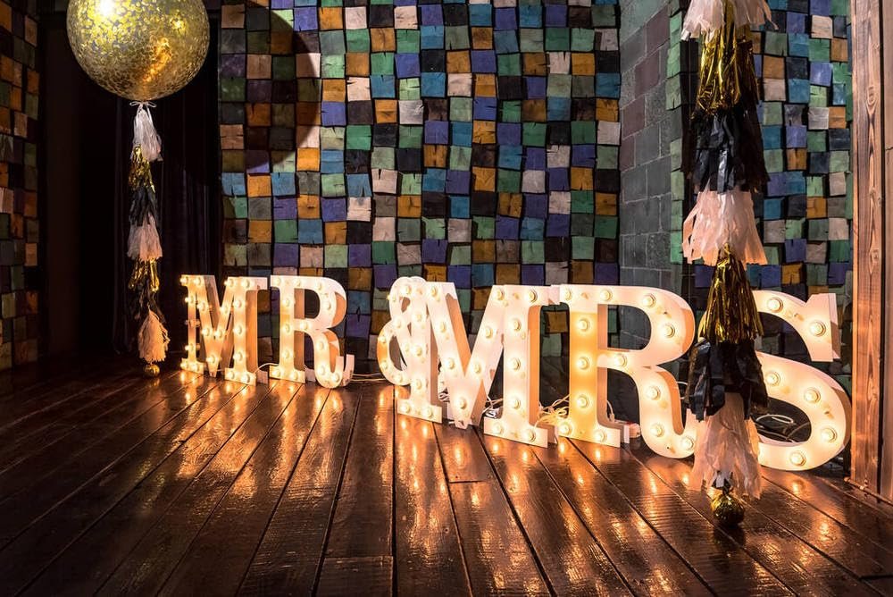 Illuminated 'MR & MRS' marquee letters on a wooden floor, with a large gold balloon and tassels.