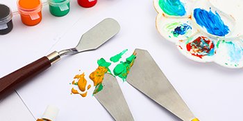 Palette knives with colorful paints on a white surface, with a paint palette and small paint pots in the background.