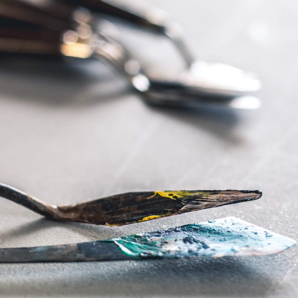 Close-up of two palette knives with paint residue, one with yellow and black paint, the other with blue and green paint.