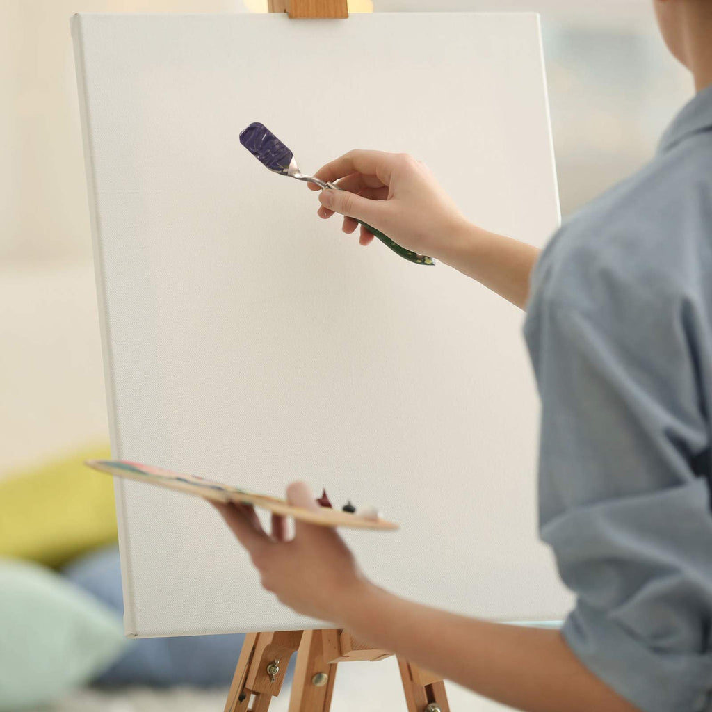 Artist's hands holding a palette knife with purple paint and a wooden palette with paints, applying paint to a canvas on an easel.