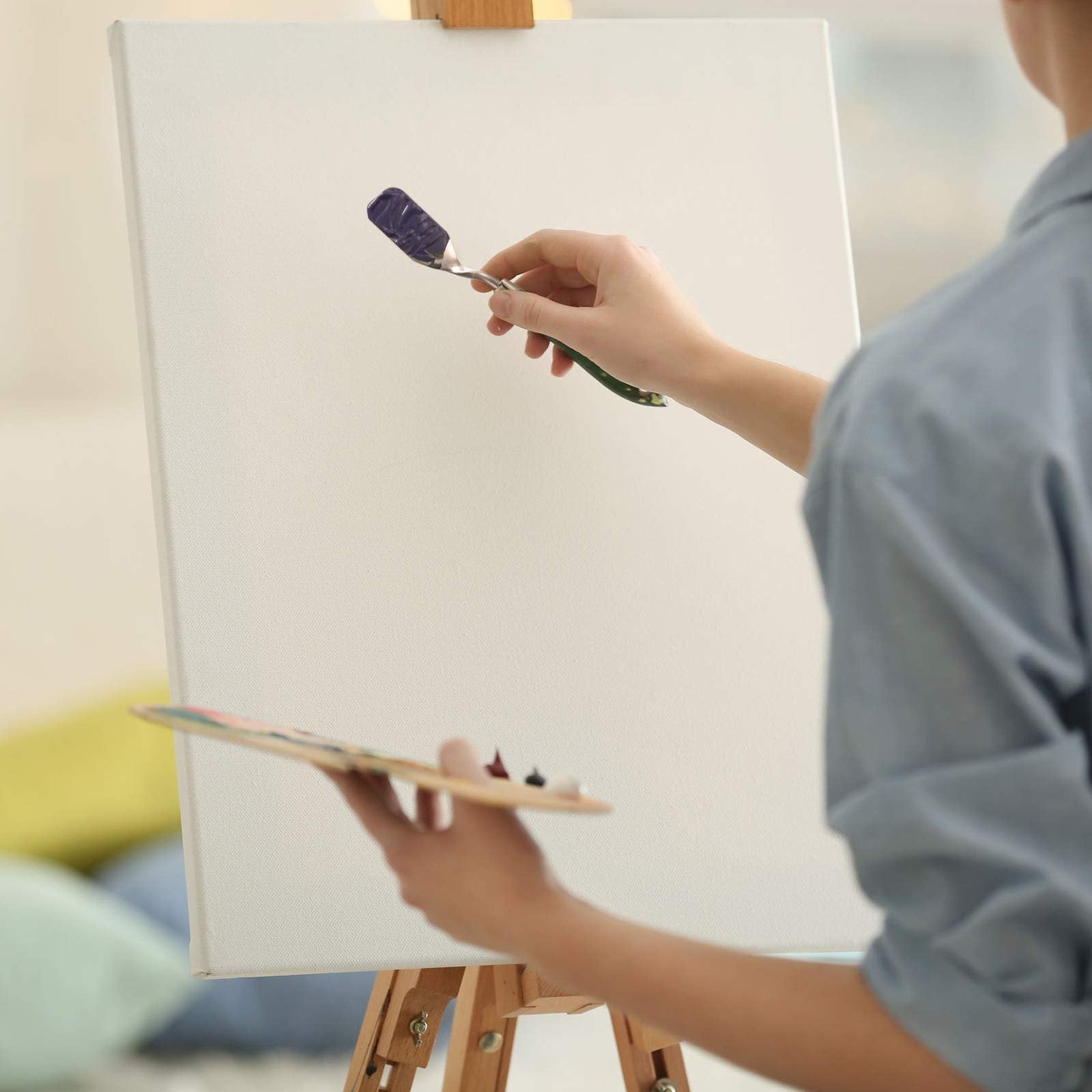 Artist's hands holding a palette knife with purple paint and a wooden palette with paints, applying paint to a canvas on an easel.