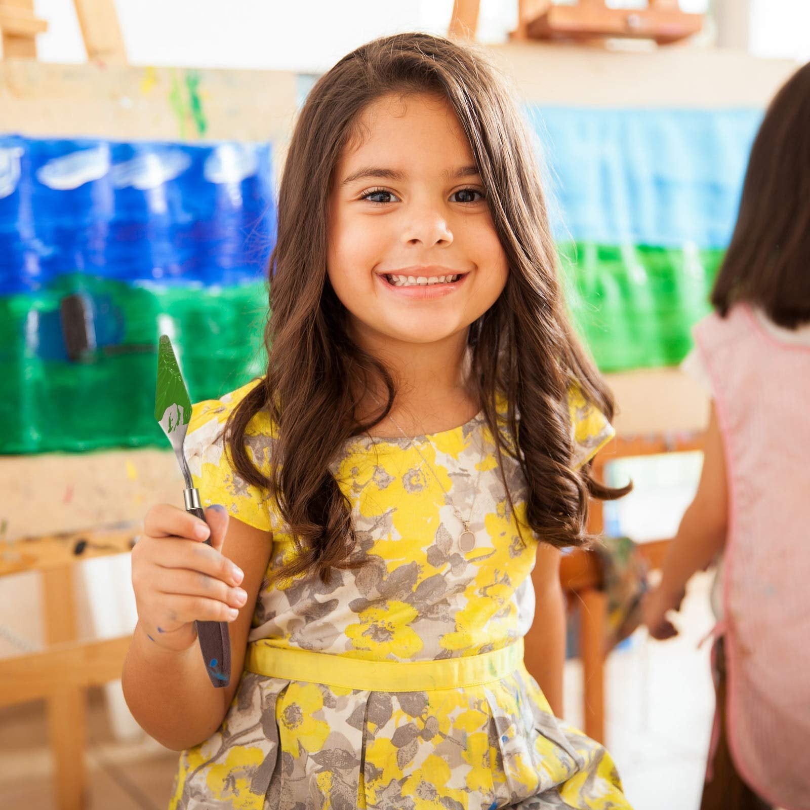 Smiling young girl holding a palette knife in front of a colorful painting, with another child in the background.
