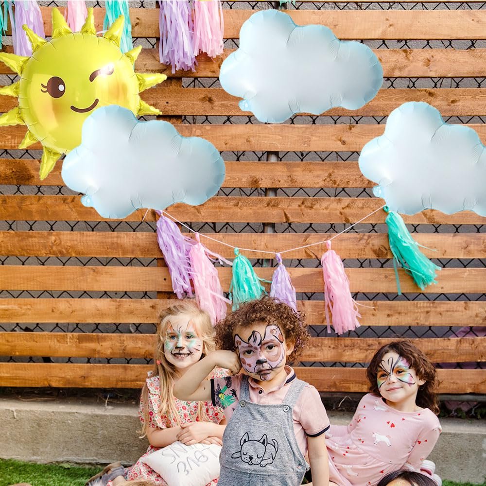 Three children with face paint sit in front of a wooden fence decorated with sun and cloud balloons and colorful tassels.