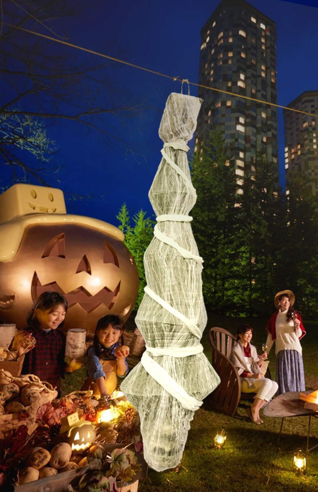 Hanging Halloween skeleton prop draped in white gauze and ribbon, outdoors at night with people and a large jack-o'-lantern.