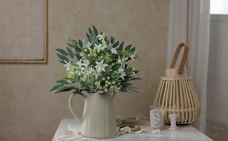 Artificial greenery stems with white flowers in a cream pitcher on a table with a wooden lantern and candle.